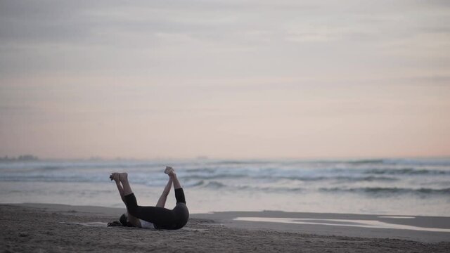 Woman At The Beach Practicing Yoga In Ananda Balasana (Happy Baby) Pose - wide shot