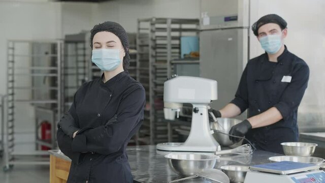 Portrait of cheerful young slim woman in Covid-19 face mask and cook uniform looking at camera smiling with blurred man posing at background. Chef in commercial kitchen in candy store on pandemic.
