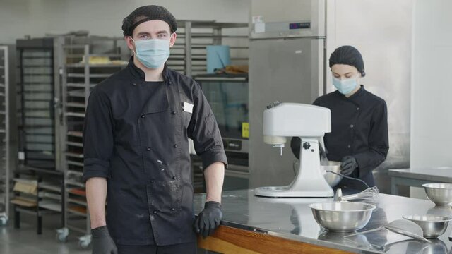 Confident man in coronavirus face mask and cook uniform turning looking at camera with blurred concentrated woman mixing dough ingredients at background. Young confectioners in kitchen on pandemic.