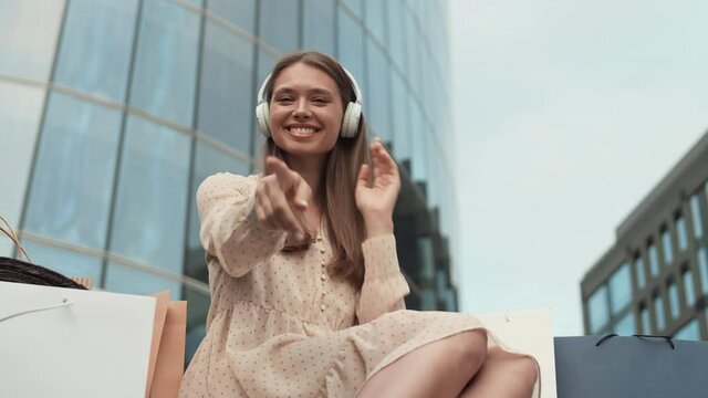 Low angle medium long POV of young Caucasian woman wearing wireless over-ear headphones, enjoying music, sitting in foreground of modern buildings, smiling, beckoning with finger, looking on camera