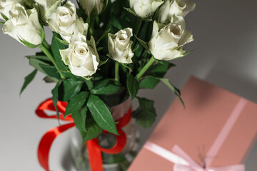 White tea roses stand in a glass vase, which is tied with a red ribbon and stands on the table. There is a gift nearby. in a pink box. Gift bouquet