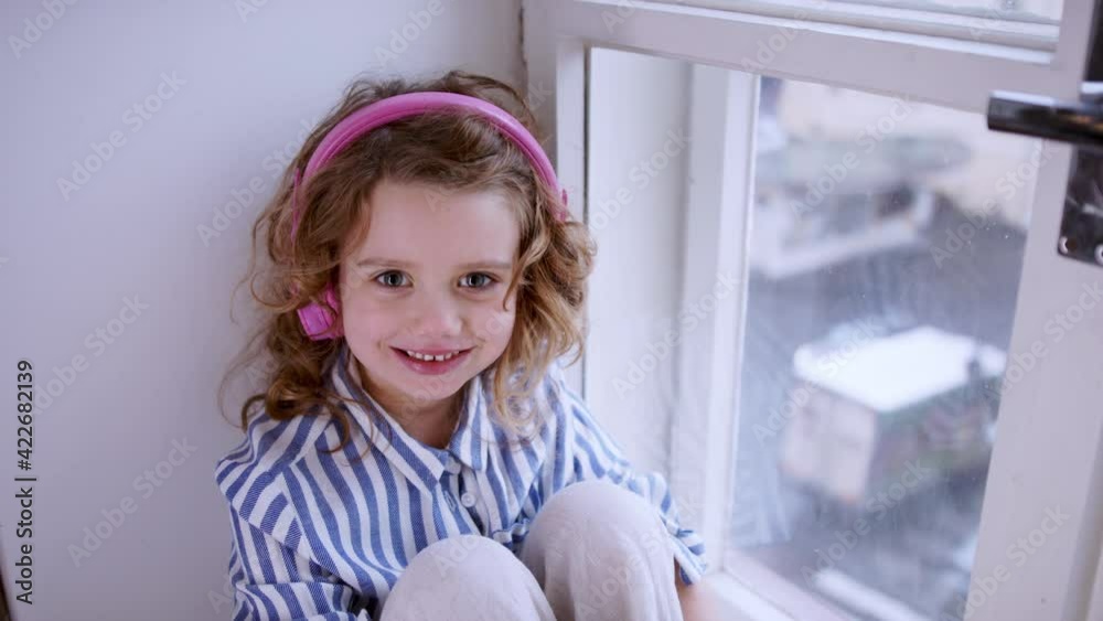 Wall mural Sad small girl with headphones indoors at home, sitting on window sill.