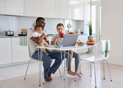 Young Black parent dad having morning breakfast with mixed race kids using laptop. Multiethnic family father and two children enjoying watching videos on computer sitting at kitchen table at home.