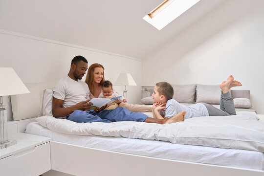Happy multiethnic family with kids having fun reading book waking up in bed together. Smiling parents African dad and Caucasian mom playing relaxing with mixed race children in bedroom at home.