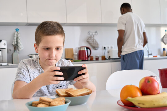 Funny Caucasian teen boy playing game on smart phone while Black African father making breakfast in the kitchen. Teenage son using mobile apps, watching videos spending time with dad at home.