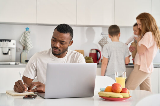 Young Black dad working or learning from home office using laptop computer while mixed race multiethnic family with diverse kids spending time in kitchen at home. Distance work and parenthood concept
