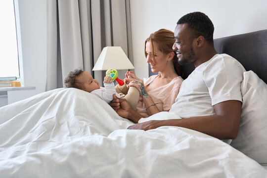 Happy mixed race young family couple lying in bed holding cute infant girl. Multiracial diverse parents mom and dad relaxing waking up in the morning playing with cute baby daughter in bedroom at home
