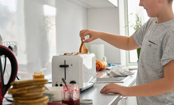 Close up view of teenage boy cooking in the kitchen, holding bread slice in hand making toasts in toaster for snack preparing family breakfast or lunch alone at home.