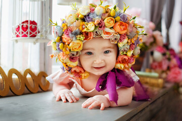 A beautiful baby girl of 6 months in a hat made of flowers, lying on her stomach by the window. A small child among the flowers with a wreath on his head.
