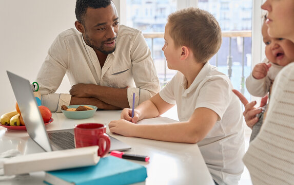 Happy teenage Caucasian school boy distance learning sitting at table with multiracial family. Black parent father helping teen son remote studying at home online using laptop homeschooling together.