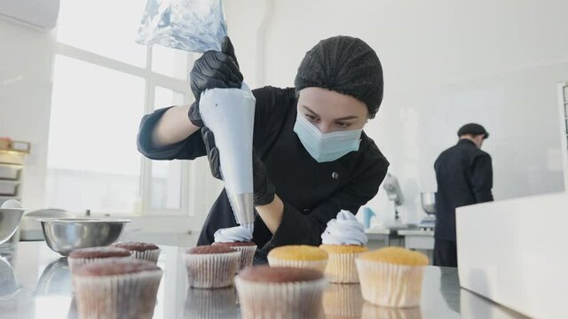 Confident professional confectioner decorating muffins with blue topping. Portrait of concentrated Caucasian woman in coronavirus face mask working in candy store kitchen indoors. Culinary and job.