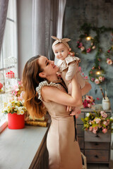 A beautiful slender mother in a beige dress holds her little daughter in her arms in the studio against the background of a vase with flowers. A woman and a baby in a spring decoration.