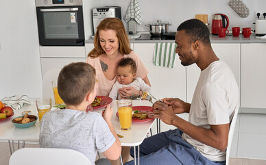 Happy multiethnic diverse family or four having morning breakfast together. Smiling couple with children eating tasty homemade pancakes enjoying talking at home sitting at modern kitchen table.