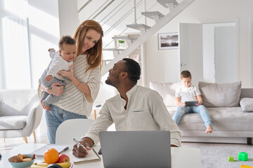 Happy African Black young father playing with cute baby child daughter while working from home office on laptop spending time with multiethnic family diverse kids and Caucasian wife at home.