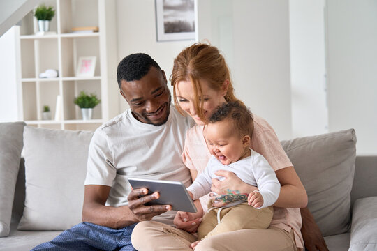 Happy young multiracial family couple holding cute infant daughter having fun with digital tablet sitting on sofa. Funny mixed race child baby girl laughing using computer with diverse parents at home