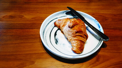Croissant with icing sugar with stainless steel knife in white dish on wooden table with copy space. Crispy bread in dish on wood background. Food for eat with coffee or tea in morning at cafe shop.