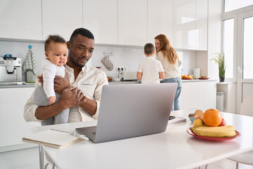 Young Black parent dad working from home office, African American father holding baby child daughter using laptop computer spending time with diverse multiracial family at home in the kitchen.