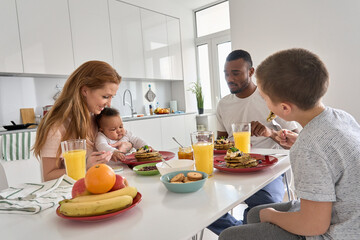 Happy multiracial diverse family couple with two children having morning breakfast at home together. Multiethnic young parents with mixed race kids eating pancakes meal sitting at kitchen table.