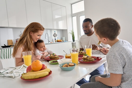 Happy Multiracial Diverse Family Couple With Two Children Having Morning Breakfast At Home Together. Multiethnic Young Parents With Mixed Race Kids Eating Pancakes Meal Sitting At Kitchen Table.