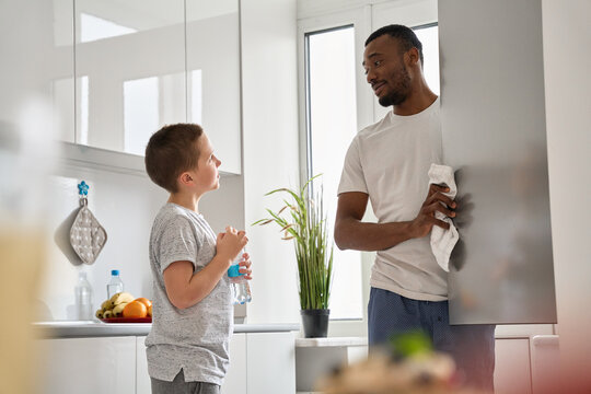 Mixed Race Family Teenage Caucasian Boy Son And Young African American Black Father Talking Standing In The Kitchen Opening Refrigerator Cooking Together Helping Doing Household Chores At Home.