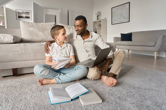 Caring Black dad hugging white teenage son helping school boy with studies. African father doing homework and teen boy virtual studying, distance learning at home with parent together sitting on floor