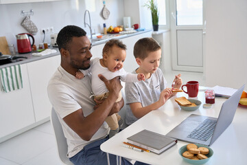 Young Black parent dad working from home office using laptop having diverse family morning breakfast with kids at home. Happy mixed race children sitting at kitchen table with African father at home.