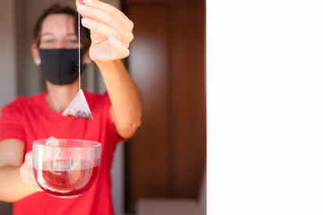 Woman with red dress is soaking tea bag on crystal cup, preparing hot tea. Dipping tea bag on cup. Woman during quarantine. There is a wall white for to write.