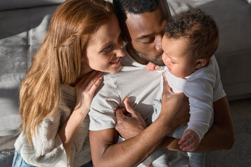 Mixed race young family couple bonding holding cute infant girl. Happy multiracial parents playing with small adorable baby daughter kissing kid at home lit with sunlight, Multiethnic family portrait