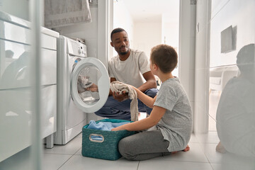 Diverse teen son and African dad doing laundry together at home. Black father and white teenage boy...