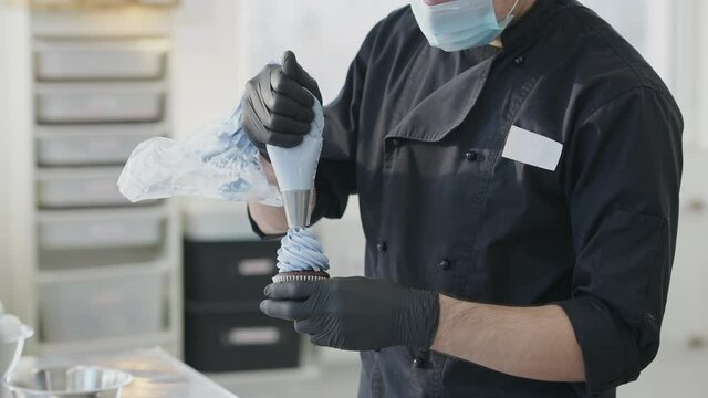 Unrecognizable concentrated Caucasian man in Covid-19 face mask pouring blue dessert topping on baked muffin. Professional male confectioner decorating pastry in candy store kitchen.