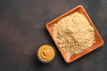 Dry mustard powder in a wooden plate and ready-made pasty mustard on a brown background....