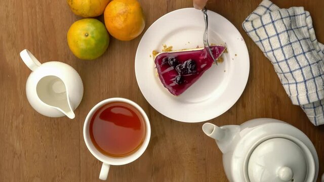 Top view of taking a bite of blueberry cheese pie in white ceramic plate with a white cup of tea and ripe oranges on wooden table