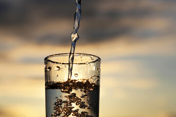 Pouring clean water into drinking glass on sunset blurred background. Concept of health and...