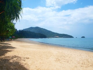 Tropical beach in Thailand. Phuket island. White sand, calm sea, blue sky with clouds. Boats in the distance. Green hills on a horizon. Palm trees and shadows from them in left side of frame. Seascape