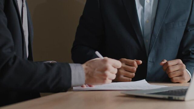 Unrecognizable Caucasian businessman and African-American man investor signing contract make handshake sitting at office desk. Close up male hands with paper documents shake hands signing agreement
