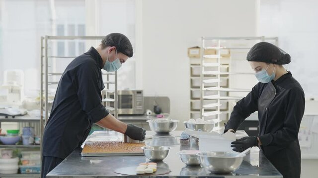 Side view of two concentrated confectioners working in candy store kitchen in slow motion. Young Caucasian man and woman cooking muffins and cakes indoors on coronavirus pandemic outbreak.