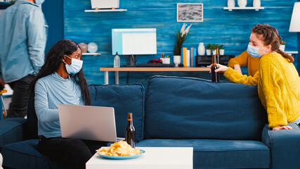 Cheerful woman holding beer bottle looking at laptop discussing with african friend in living room wearing face mask to prevent coronavirus spread in time of global pandemic. Conceptual footage.