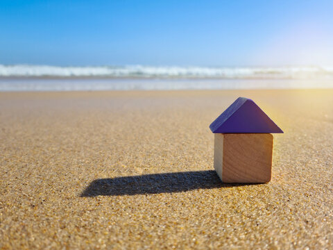 Colourful blue yellow red miniature wooden kids toy home model on sunny sand beach with blue sky, ocean background. Copy space of family lifestyle and business real estate property investment concept
