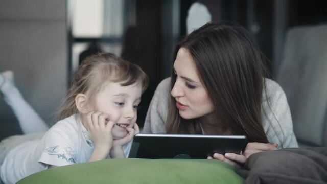 Cute Little Small Kid Child Daughter With Adult Mum Using Digital Tablet Technology Device Surfing Internet Using Education Apps Touching An Electronic Pad Screen Smiling Laying On Sofa At Home