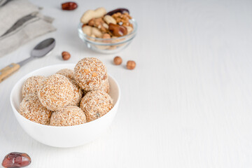 Organic vegetarian truffles or homemade nut vegan energy balls made of sesame, hazelnut and dates fruit served in bowl with spoon on white wooden table with textile. Image with copy space, horizontal