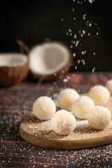 Coconut organic sweet truffles or homemade nut vegan energy balls with sprinkles of shredded coconut  served on cutting board on dark brown wooden table at kitchen against black wall. Vertical
