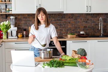 Close up woman in white t-shirt cooking soup with fresh vegetables in kitchen at home. Menu, recipe book banner. Girl reads the recipe in laptop. Caucasian model using internet in loft apartment.
