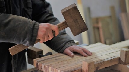 a carpenter in dark dusty work clothes holds a wooden hammer over a piece of glued wooden planks on a work table in a workshop, the process of using a mallet in carpentry