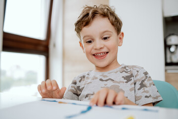 Naklejka premium Portrait of a lovely happy school-aged boy sitting at kitchen table enjoying some free time. Little boy is making something out of plasticine.