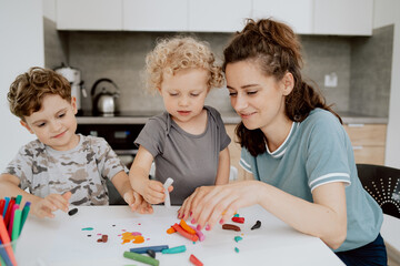 A young pretty mother is sitting at the kitchen table with her preschool-aged daughter and school-aged son making something out of plasticine. The mother is spending free time with her children.