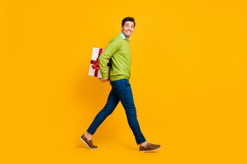 Full length body size profile side view of nice funky cheerful guy carrying hiding giftbox isolated on bright yellow color background