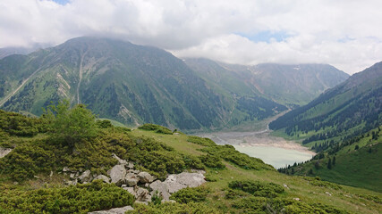 Fototapeta na wymiar Big Almaty lake in the mountains. Green hills, flowers and fields. White clouds over the mountains. Mountain lake. In places there are stones. Protected place of nature, reserve of TRANS-ili Alatau