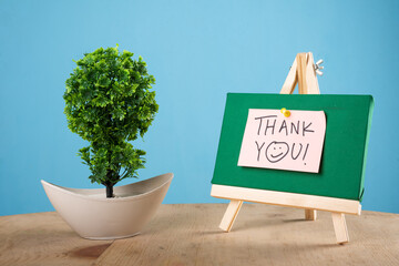 Thank you written on a sticky note on a mini green notice board with a plant with blue background