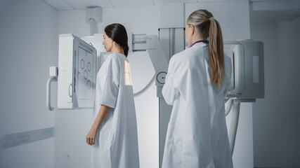 Hospital Radiology Room: Beautiful Multiethnic Woman in Medical Gown Standing Next to X-Ray Machine while Female Doctor Adjusts it. Healthy Patient Undergoes Routine Scanning with the Nurse's Help.