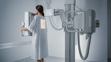 Hospital Radiology Room: Beautiful Multiethnic Woman Standing in Medical Gown in the X-Ray Machine. Adult Female Undergoes Healthcare Exam and is Scanning Chest, Heart, Lungs in Modern Clinic Office.
